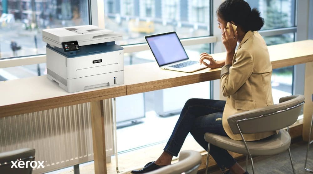 Xerox® Print and Scan Experience: Convenient and Connected A person is sitting at a high table by a large window, talking on a phone while working on a laptop. A Xerox B235DNI multifunction printer is conveniently placed on the table next to them, suggesting ease of use in a modern workspace.